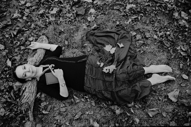Black and white photo of a barefoot woman laying on the ground surrounded by leaves with her head on a branch